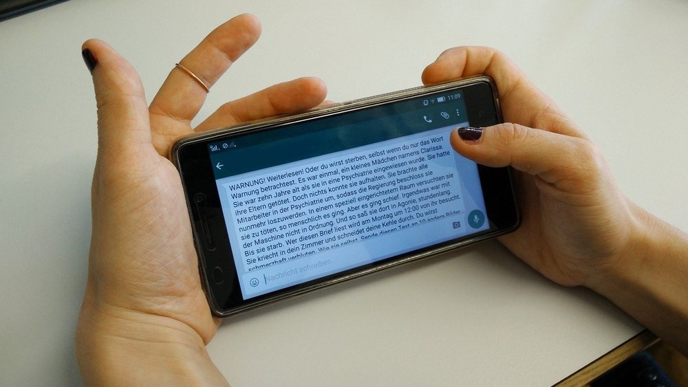 Nahaufnahme von zwei Händen, die ein Smartphone halten. Am Display ist WhatsApp mit einer Kettenbrief-Nachricht zu sehen.