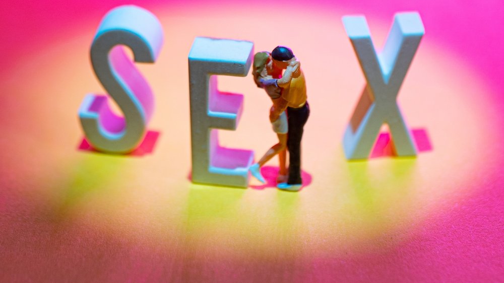 Zwei Figuren küssen sich zwischen den Buchstaben E und X, insgesamt ist das Wort SEX zu lesen