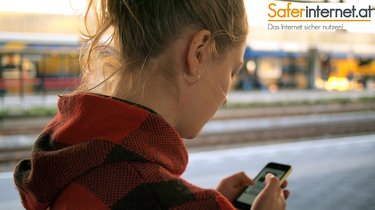 Eine junge Frau nutzt auf einem Bahnsteig ein Handy und ist von hinten von der Kamera aufgenommen worden