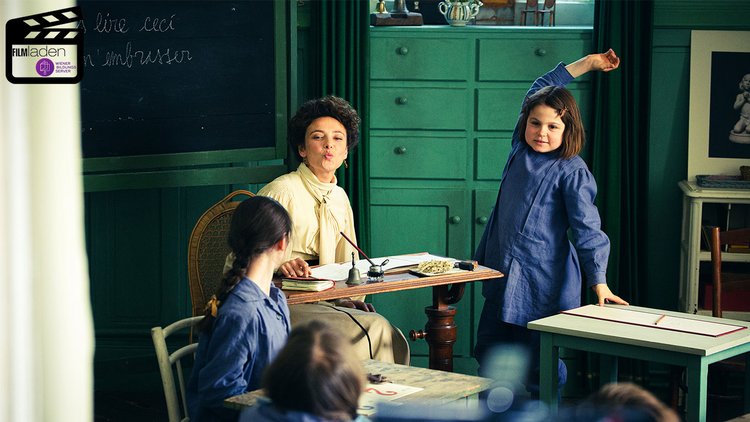 Maria Montessori sitzt an einem Lehrerpult in einem Klassenzimmer vor einer Tafel und lächelt, einen Kussmund formend, andere Schüler:innen an. Neben ihr zeigt eine Schülerin auf.