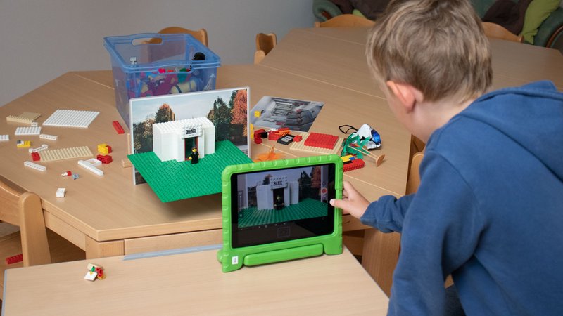 Ein Junge lehnt sich am Tisch zu einem Tablet, vor dem Tablet ist vor einem Hintergrundbild eine Spielfigur aufgebaut.