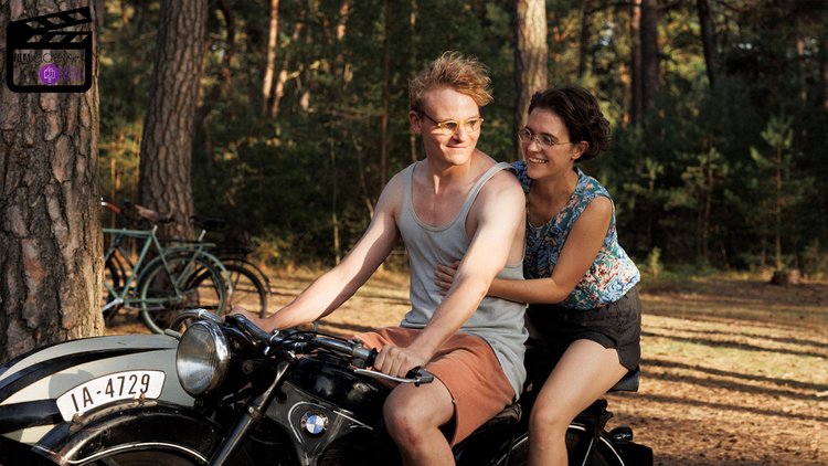 Ein Mann fährt ein Motorrad und lächelt. Hinter ihm am Motorrad sitzt eine Frau, die sich an ihm festhält und ihn anlächelt.