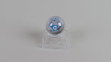 Der Sphero Mini Roboter auf einer transparenten Halterung vor grauem Hintergrund.