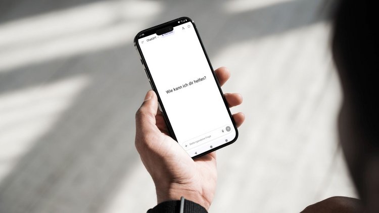 Nahaufnahme einer Person, die ein Smartphone in der Hand hält. Am Bildschirm ist die Startoberfläche von ChatGPT zu sehen mit der Frage "Wie kann ich dir helfen?".