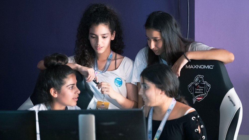 Eine Gruppe jugendlicher Mädchen sitzen und stehen hinter zwei Computerbildschirmen, sie sprechen miteinander