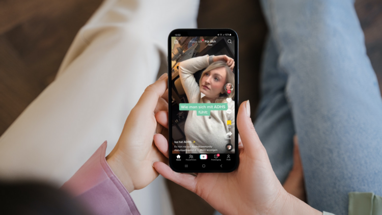 Eine Person sitzt auf einer Couch und hält ein Smartphone in den Händen. Am Display ist ein TikTok-Video mit dem Titel "Wie man sich mit ADHS fühlt" zu sehen.