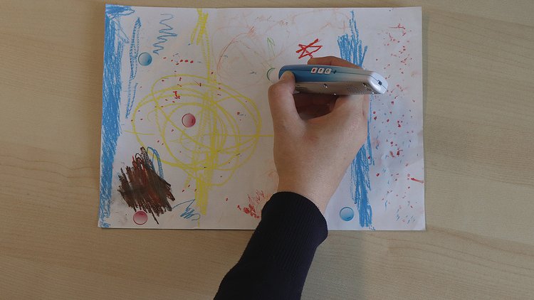 Auf einem Tisch liegt ein Blatt mit Zeichnung, eine Hand mit Audiostift berührt Audiosticker darauf
