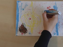 Auf einem Tisch liegt ein Blatt mit Zeichnung, eine Hand mit Audiostift berührt Audiosticker darauf