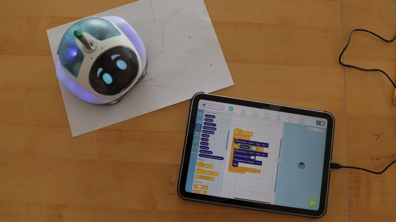 Der Loti-Bot zeichnet mit einem Stift in der rundlichen Öffnung auf einem Blatt Papier Linien. Daneben liegt ein Tablet, darauf ist die Scratch-Programmieroberfläche in der App zu sehen.