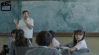 Foto eines Lehrers, der gestikulierend vor einer Schultafel steht, auf der einige Bruchrechnungen aufgeschrieben sind. Vor ihm sitzen mehrere Schülerinnen und Schüler.