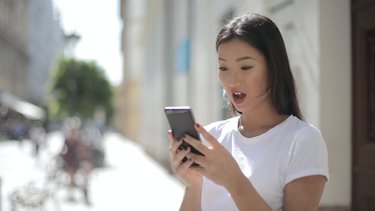 Eine junge Frau hält ein Smartphone in ihren Händen und schaut mit schockiertem Gesichtsausdruck auf das Smartphone.