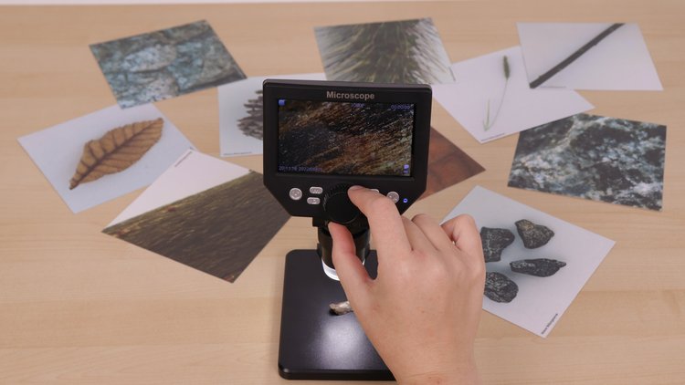 Ein Digitales Mikroskop steht auf einem Tisch. Daneben liegen Bildkarten, auf denen verschiedene Naturmaterialien zu erkennen sind. Eine Hand tippt auf das Digitale Mikroskop.