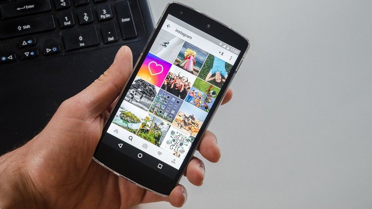 Nahaufnahme einer Hand, die ein Smartphone hält. Auf dem Display des Smartphones ist ein geöffnetes Instagram-Profil zu erkennen. Im Hintergrund ist eine Computertastatur zu sehen. 