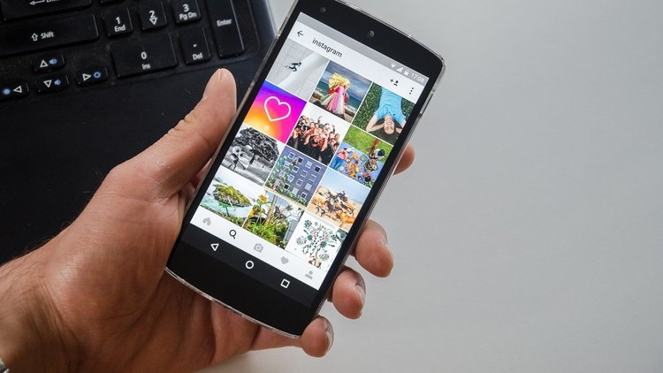 Nahaufnahme einer Hand, die ein Smartphone hält. Auf dem Display des Smartphones ist ein geöffnetes Instagram-Profil zu erkennen. Im Hintergrund ist eine Computertastatur zu sehen. 