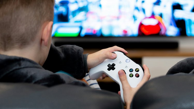 Ein Junge sitzt mit einem Konsolencontroller auf einem Sofa und spielt ein Spiel auf einem großen Bildschirm