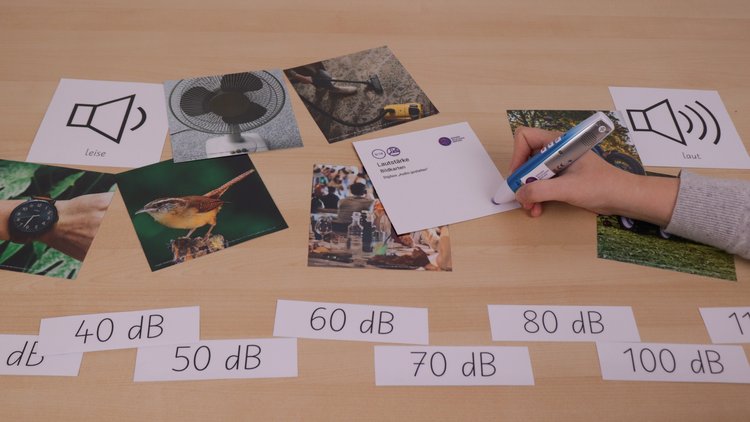 Auf einem Tisch liegen verschiedene Bildkarten. Eine Hand hält einen Tellimero-Stift und tippt auf einen bunten Sticker.