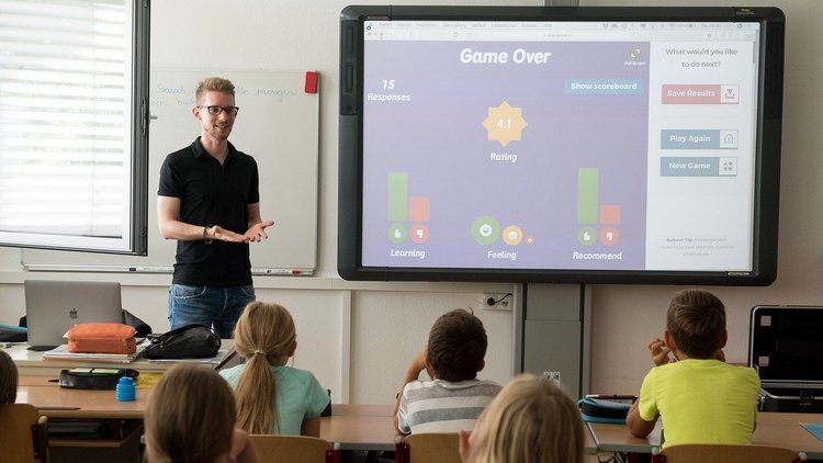 Foto in einem Klassenraum: Schüler und Schülerinnen der Primarstufe sitzen an Schreibtischen, vorne steht neben einem Interactive Whiteboard ein Lehrer. Am Whiteboard ist die Oberfläche von Kahoot zu sehen.