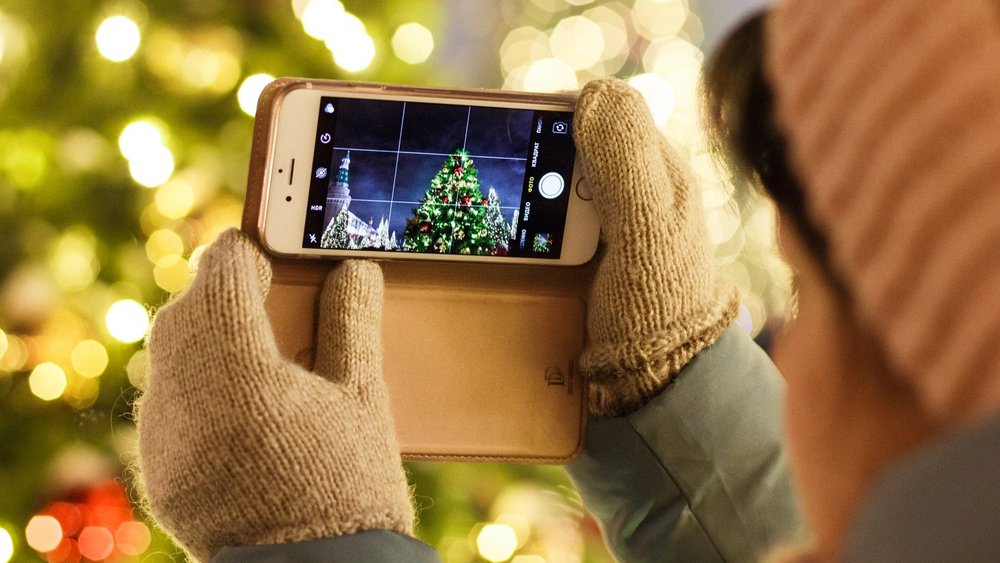 Nahaufnahme über die Schulter einer Person, die ein Smartphone quer hält und damit einen großen Weihnachtsbaum fotografiert.