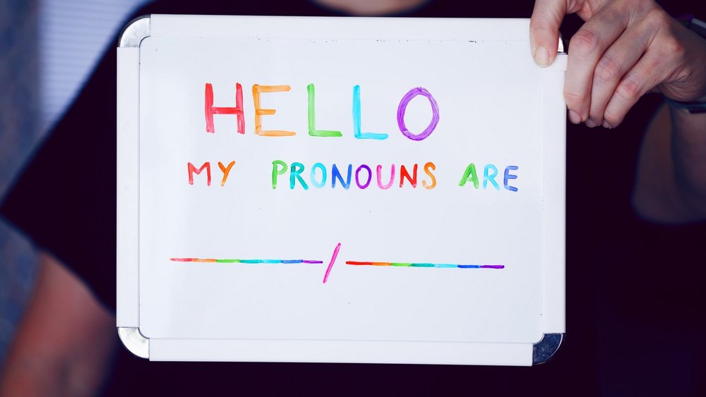 Eine Person hält ein Schild in die Kamera, darauf steht geschrieben "Hello My pronouns are ___ / ___"