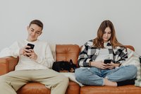 Ein männlicher und ein weiblicher Teenager sitzen nebeneinander auf einem braunen Ledersofa, dazwischen liegt schlafend ein kleiner, schwarzer Hund. Beide Jugendliche schauen auf Smartphones, die sie in ihren Händen halten.