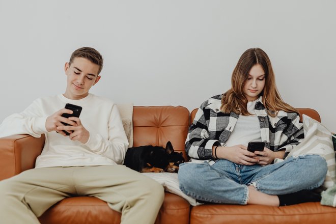 Ein männlicher und ein weiblicher Teenager sitzen nebeneinander auf einem braunen Ledersofa, dazwischen liegt schlafend ein kleiner, schwarzer Hund. Beide Jugendliche schauen auf Smartphones, die sie in ihren Händen halten.