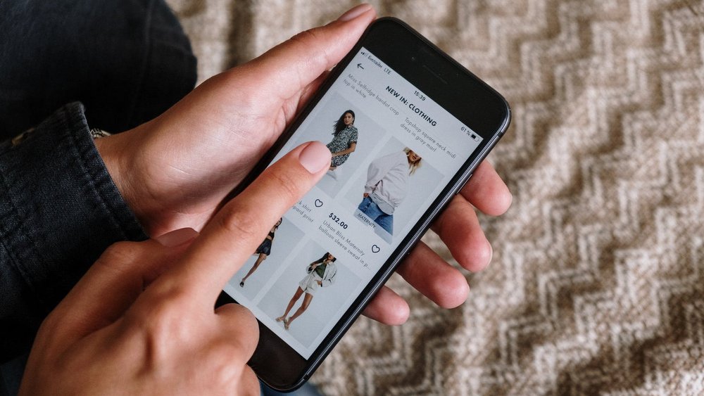Nahaufnahme: Eine Hand hält ein Smartphone, am Display ist eine Online-Shopping-App mit Kleidung zu sehen. Eine zweite Hand tippt auf ein Kleidungsstück.