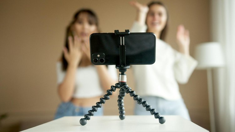 Nahaufnahme eines Smartphones, das auf einem kleinen Stativ steht. Hinter dem Smartphone sind unscharf zwei Jugendliche zu erkennen, die Tanzbewegungen machen.