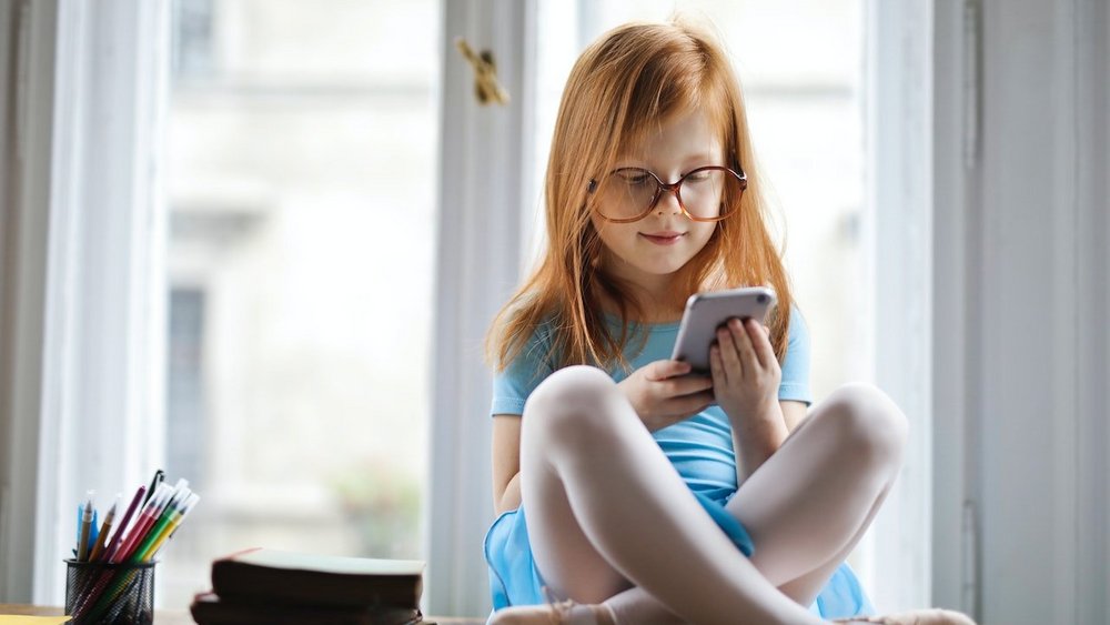 Ein junges Mädchen sitzt auf einem Fensterbrett, hält ein Smartphone in den Händen und schaut auf den Display.