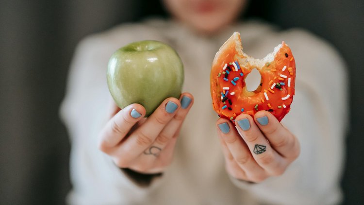 Nahaufnahme: eine Hand hält einen Apfel, eine Hand hält einen Donut. 