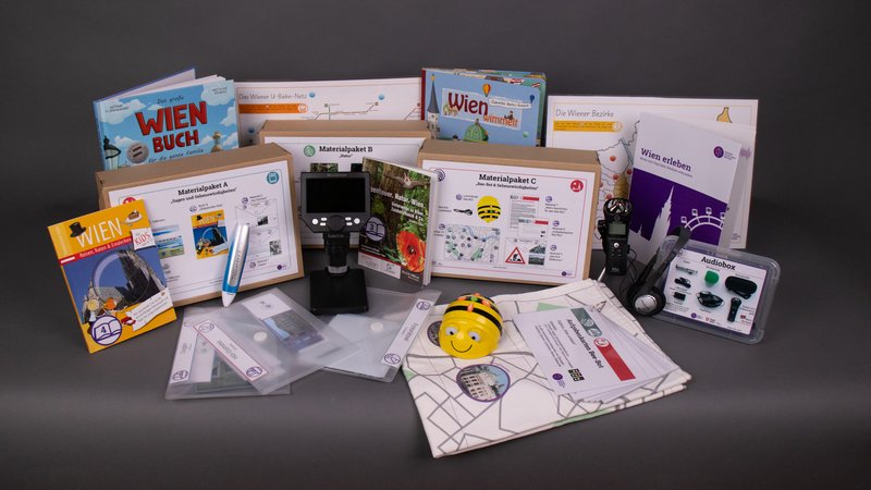 Der Inhalt der Wien erleben-Digibox: Diverse Wien-Bücher für Kinder und pädagogische Materialpakete sowie technische Geräte sind abgebildet. Darunter ein Tellimero-Audiostift, ein Bee-Bot-Roboter, ein Audioaufnahmegerät und ein digitales Mikroskop.