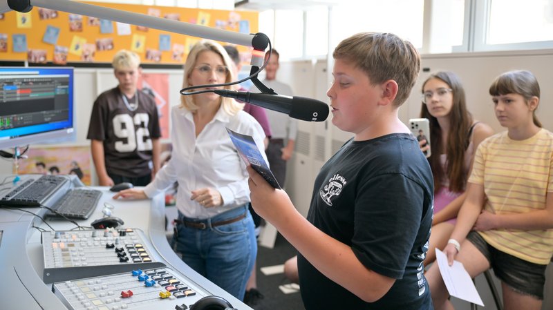 Ein Schüler spricht an einer Radio-Station in ein Mikrofon, eine Erwachsene und andere Schüler:innen stehen im Hintergrund und sehen zu.
