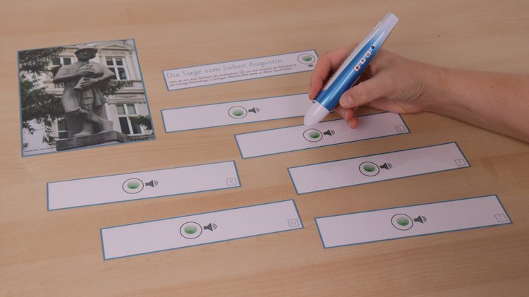 Auf einem Tisch liegen verschiedene Kärtchen und ein Bild. Eine Hand hält einen Audiostift und tippt damit auf einen Sticker auf eines der Kärtchen.