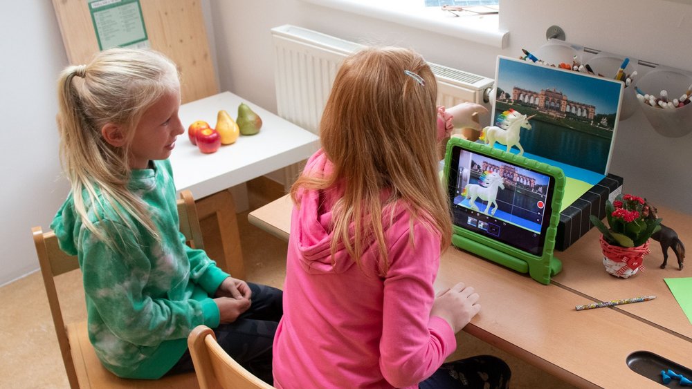 Zwei Mädchen sitzen mit einem Tablet am Tisch, vor dem Tablet bewegen sie vor einem Hintergrundbild zwei Spielfiguren.