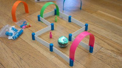 Auf einem Holzboden ist ein Parcours aus weißen Schienen und bunten Toren und Kegeln aufgebaut. Ein leuchtender Sphero Mini-Roboter bewegt sich durch den Parcours.