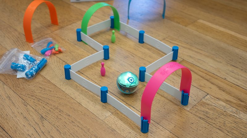 Auf einem Holzboden ist ein Parcours aus weißen Schienen und bunten Toren und Kegeln aufgebaut. Ein leuchtender Sphero Mini-Roboter bewegt sich durch den Parcours.