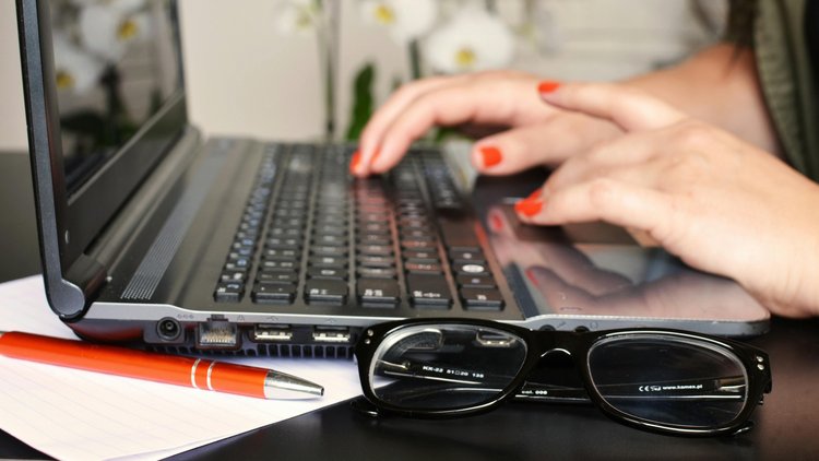 Nahaufnahme zweier Hände, die auf einer Laptop-Tastatur tippen. Vor dem Laptop liegt zusammengeklappt eine Brille.