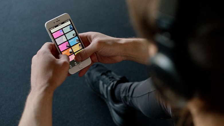 Nahaufnahme über die Schulter einer Person mit Kopfhörern, die ein Smartphone in Händen hält auf dem die App Spotify geöffnet ist. Am Smartphone-Screen werden verschiedene Musikkategorien angezeigt.