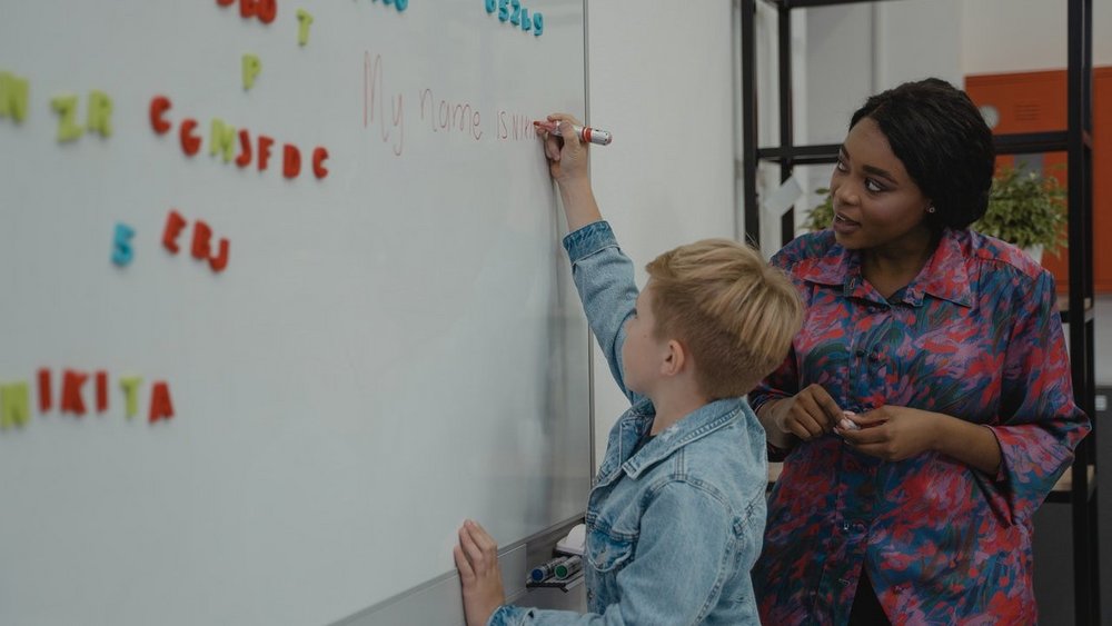 Ein Kind steht an der Tafel und schreibt darauf die Worte "My name is", daneben steht die Lehrerin und sieht zu.