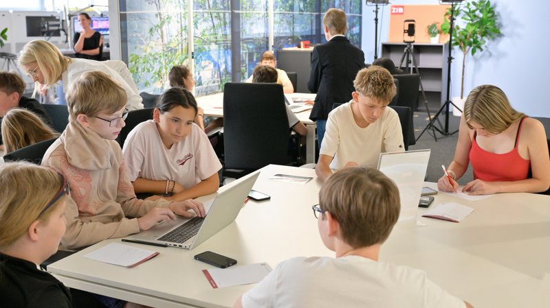Schüler:innen sitzen in Kleingruppen unterteilt an verschiedenen Tischgruppen und arbeiten mit Laptop, Stift und Papier.