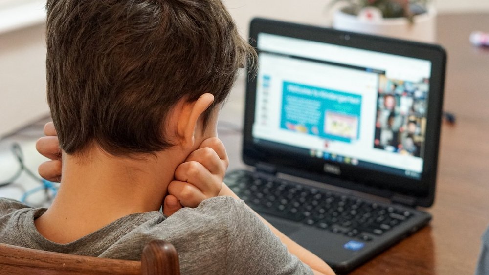 Ein Bub sitzt vor einem Laptop und hat den Kopf in seine Hände gestemmt.