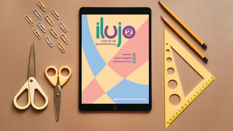 Auf einem Tisch liegen mehrere Bürogeräte und ein Tablet, auf dem das Cover von "Ilujo 2" zu sehen ist