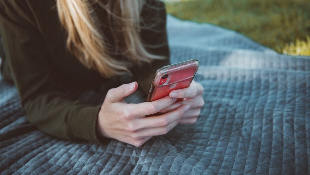 Eine junge Frau liegt in einer Wiese auf einer Decke und schaut auf ein Smartphone, dass Sie in Ihren Händen hält.