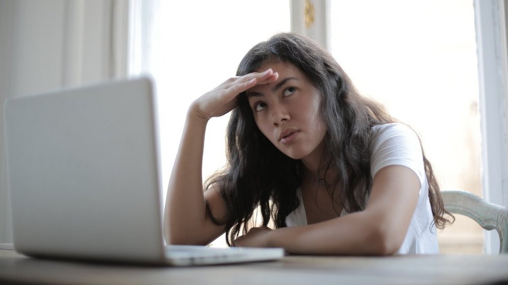 Eine junge Frau sitzt mit genervtem Gesichtsausdruck vor einem Laptop und greift sich an die Stirn.