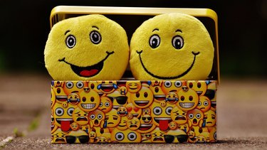 Zwei lächelnde Plüsch-Emojis schauen aus einer kleinen Box, die mit unzähligen Emojis bedruckt ist.