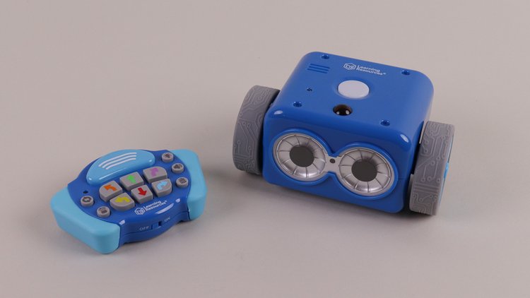 Der blaue, quadratische Botley 2.0-Roboter steht vor grauem Hintergrund. Daneben liegt die Fernbedienung, mit diversen Pfeiltasten darauf.