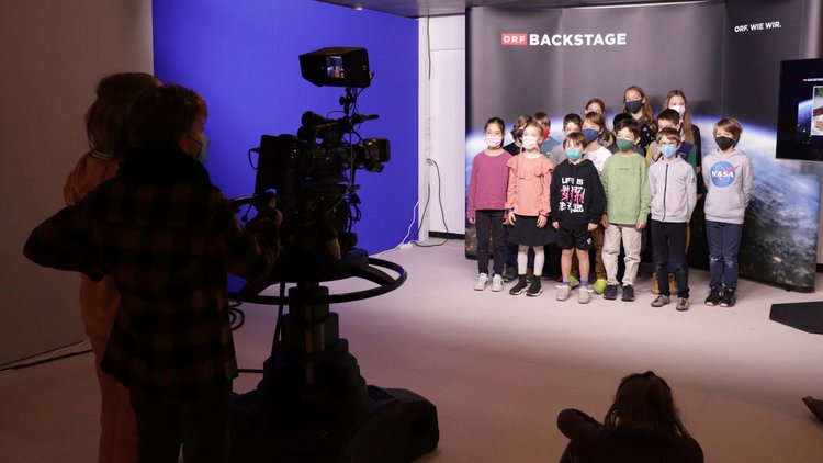 Zu sehen ist ein kleines TV-Studio: Zwei Kinder stehen im Dunklen hinter einer großen Kamera, vor einer "ORF Kids News"-Kulisse steht eine Volksschulklasse.