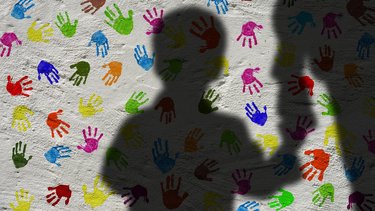 Der Schatten eines Kindes an der Hand eines Erwachsenen, an der Wand viele bunte Abdrücke von Kinderhänden