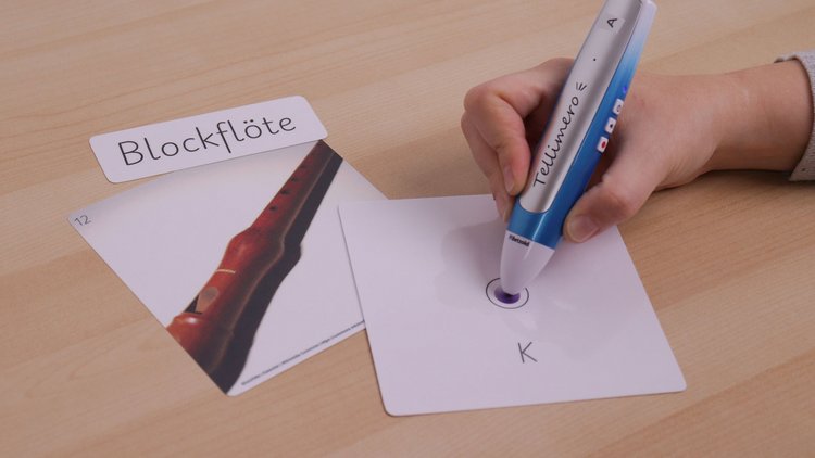 Eine Hand hält einen Tellimero-Audiostift und tippt damit auf ein Kärtchen mit einem runden, lila Sticker. Daneben liegt ein Bild von einer Blockflöte und ein Kärtchen mit dem Schriftzug "Blockflöte".