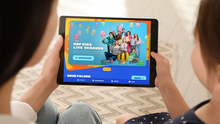 Zwei Kinder sitzen nebeneinander und halten mit je einer Hand ein Tablet, die Kamera fotografiert über die Schultern der Kinder. Am Tablet ist die ORF Kids App geöffnet.