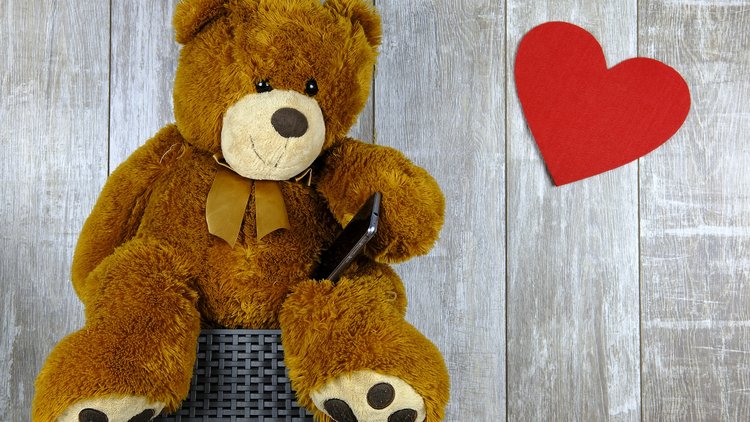 Ein Plüschbär hält ein Handy, ein Filzherz schwebt rechts neben ihm